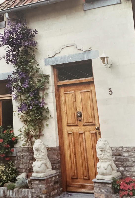 De voordeur van het huis: een houten deur met het huisnummer 5, twee stenen leeuwen en een paarse clematis