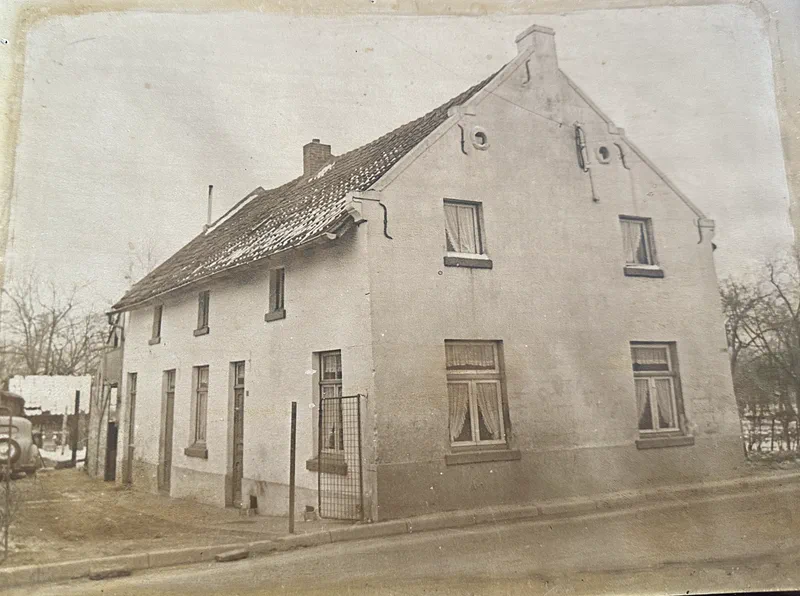 Oude zwart-witfoto: het mergelstenen huis met de puntgevel vlak langs de weg, een hekje en een auto van toen