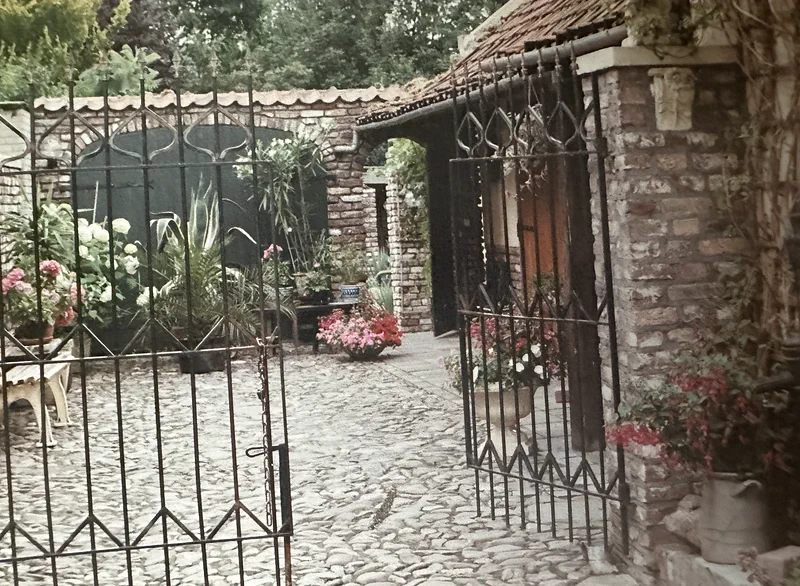 Het smeedijzeren hek staat open naar de binnenplaats met keien, planten en een schuur